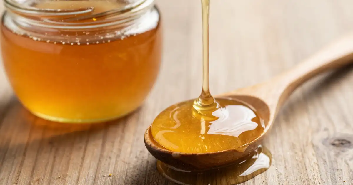 A wooden spoon full of honey gently dripping down and an open jar of honey on a wooden table.
